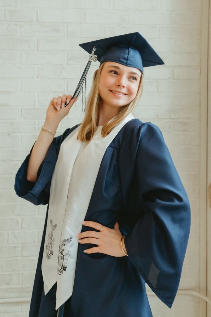 Smiling graduate in navy cap and gown holding tassel, wearing white stole indoors.