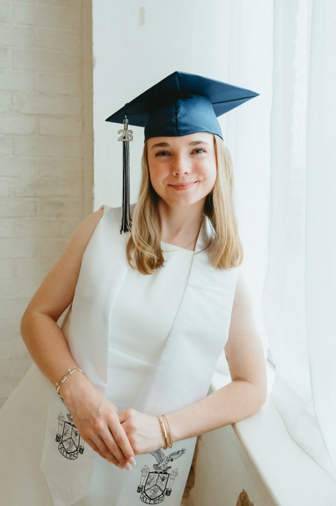 Young woman in white dress and navy graduation cap smiling near a bright window with honor stole.