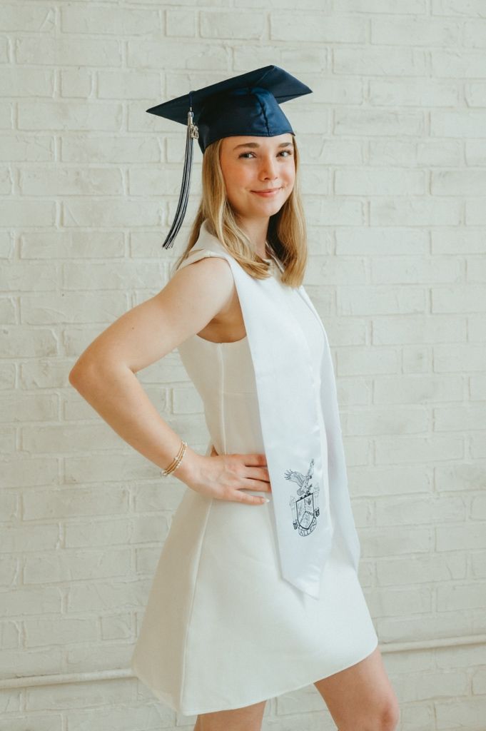 Young woman in white dress and navy graduation cap poses confidently against white brick wall.
