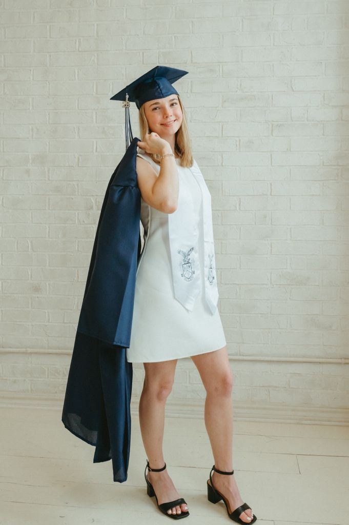 Smiling blonde graduate in white dress and navy cap holding graduation gown over shoulder.