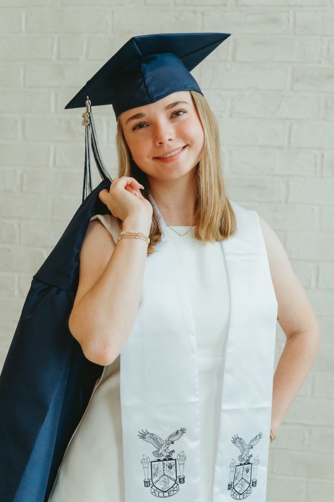 Smiling blonde graduate in navy cap holding gown with white stole featuring eagle crest emblem.