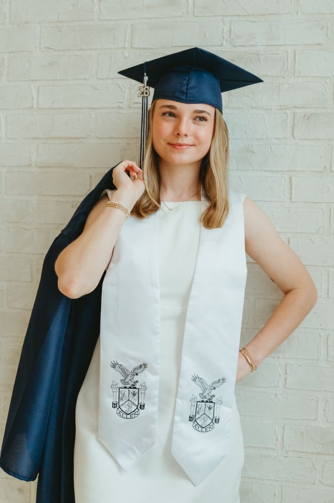 Young woman in graduation cap and white dress with stole poses proudly against a brick wall.