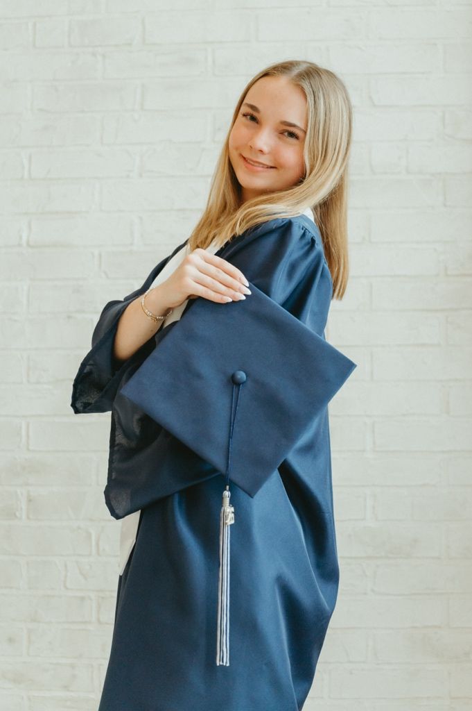 Smiling blonde graduate in navy blue cap and gown holding mortarboard against white brick wall.