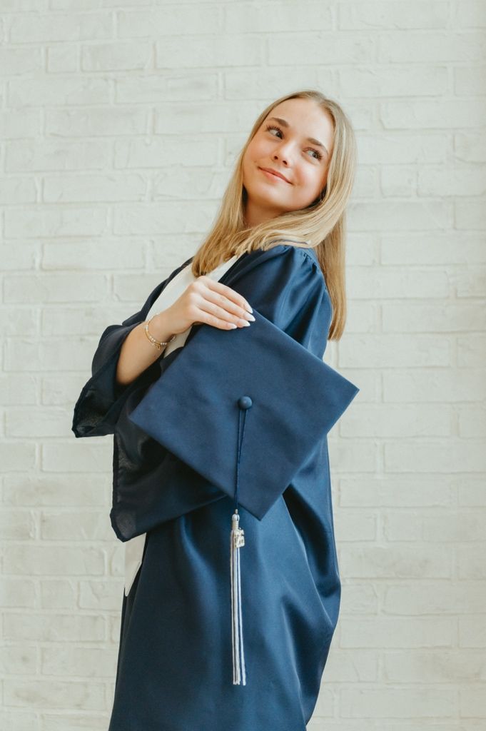 Young blonde woman in navy blue graduation gown and cap smiling against white brick wall.