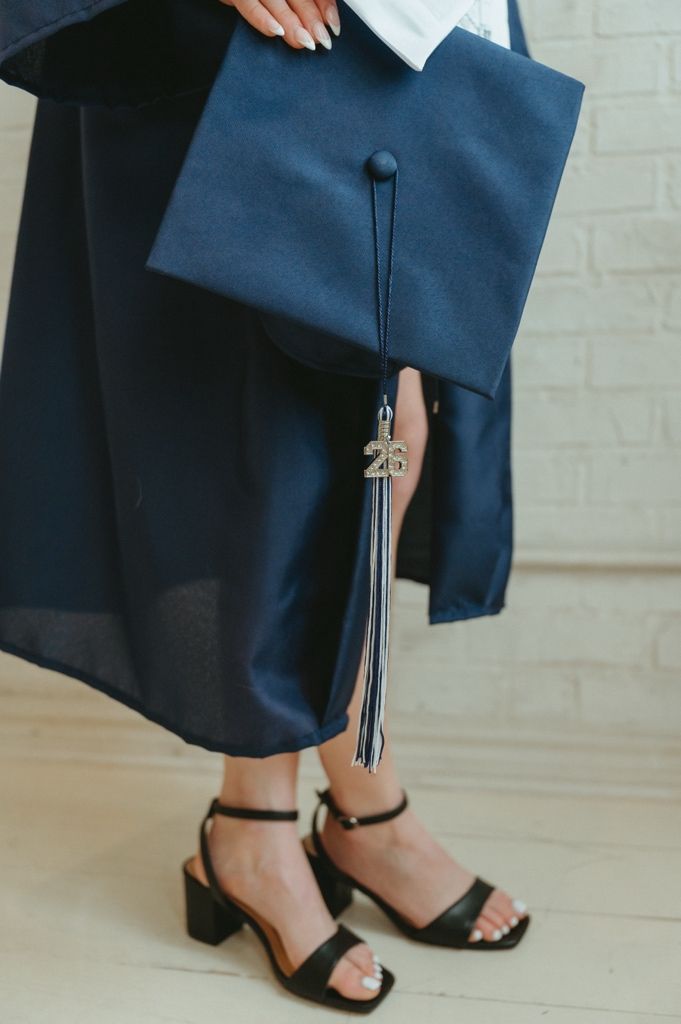 Close-up of a graduate holding a navy blue cap with a 2026 tassel, wearing black heeled sandals.