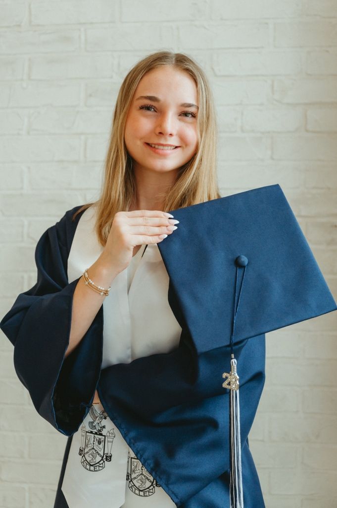Smiling blonde woman in navy graduation gown holding mortarboard cap against white brick wall.
