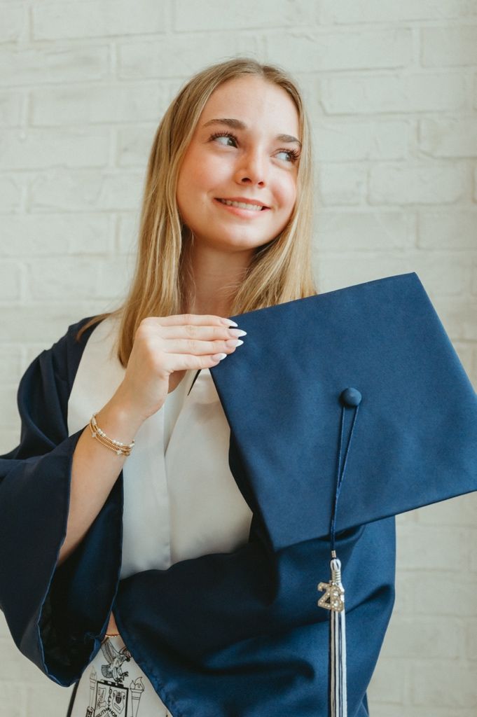 Happy blonde graduate in navy cap and gown looking up while holding mortarboard against brick wall.
