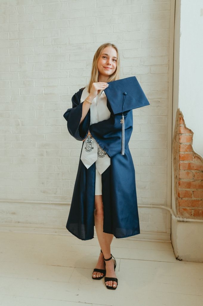 Smiling female graduate in navy cap and gown holding her graduation cap against a white brick wall.