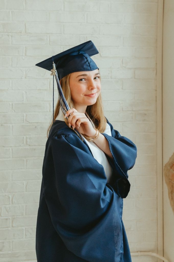 Smiling young woman in navy blue graduation cap and gown holding tassel against white brick wall.