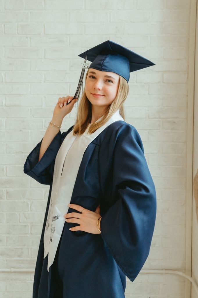 Blonde woman posing in navy graduation cap and gown with white stole against white brick wall.