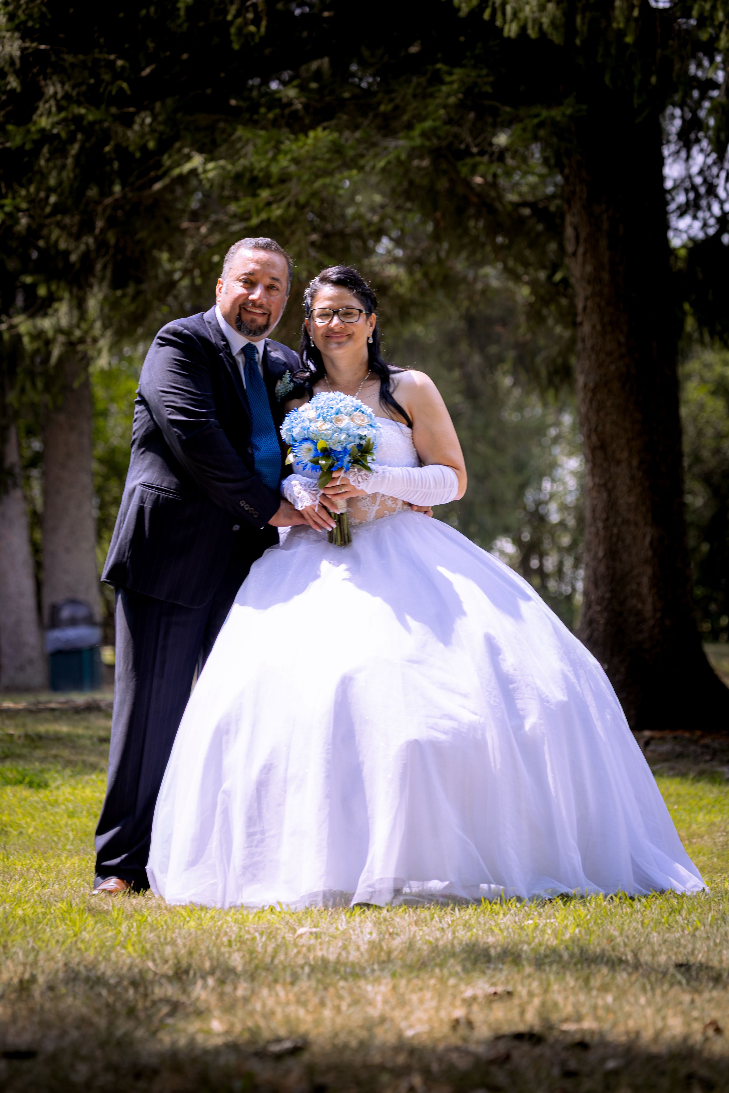 Bride in white ball gown holding blue bouquet poses with groom in dark suit outdoors in a park.