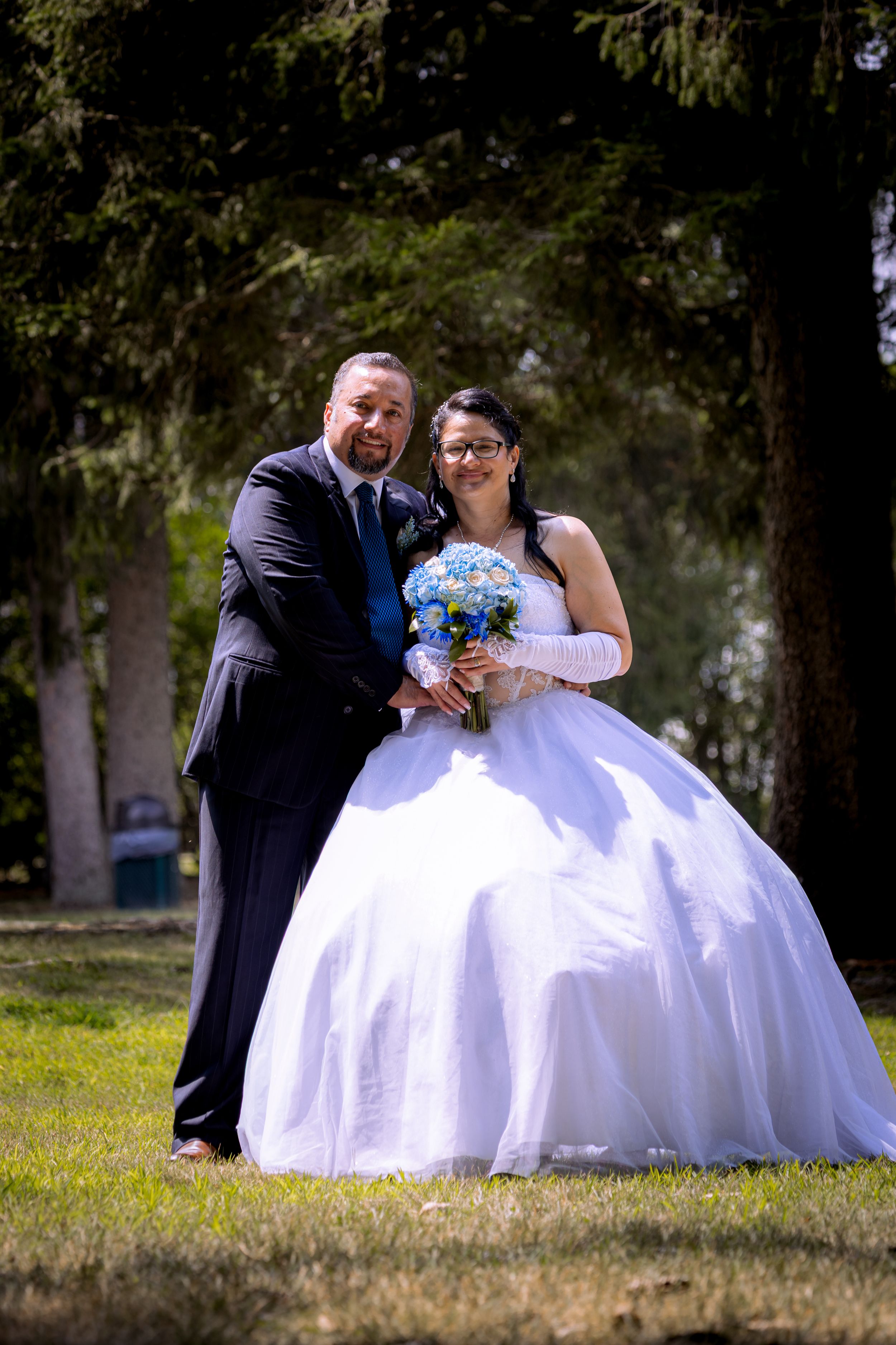 Bride in white ball gown holding blue bouquet poses with groom in dark suit outdoors in a park.