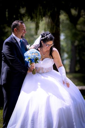 Groom in dark suit presents blue hydrangea bouquet to bride in white ball gown in a sunny park.