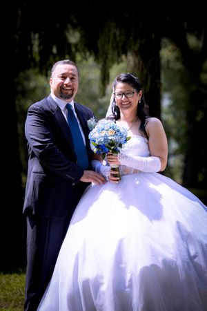A smiling bride in a white ballgown holding blue hydrangeas stands with her groom in a dark suit and blue tie outdoors.