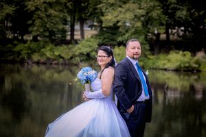 A bride in a white gown holding blue flowers stands back-to-back with a groom in a dark suit by a serene pond.