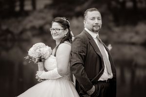 Black and white wedding photo of bride holding bouquet and groom standing back to back near a lake.