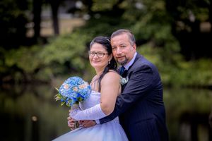 A bride and groom pose outdoors near a pond, she holds a blue bouquet in a white gown, he wears a dark suit.