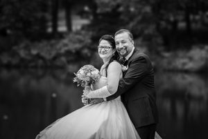 Black and white wedding photo of smiling bride holding bouquet and groom embracing near a lake.