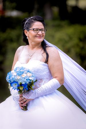 Bride in white ball gown with lace gloves holds blue and white bouquet, smiling outdoors.