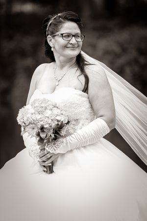 Black and white portrait of a smiling bride in strapless gown, veil, glasses, gloves, holding a bouquet.
