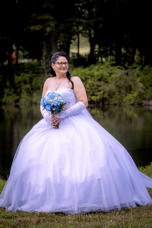 Elegant bride in voluminous white tulle gown holds blue bouquet near serene pond backdrop.