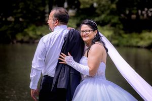 Bride in white gown and veil embraces groom in dark suit outdoors near a pond surrounded by lush green trees.