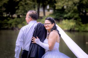Bride in white gown and veil embraces groom in black suit near a serene lake surrounded by lush green trees.
