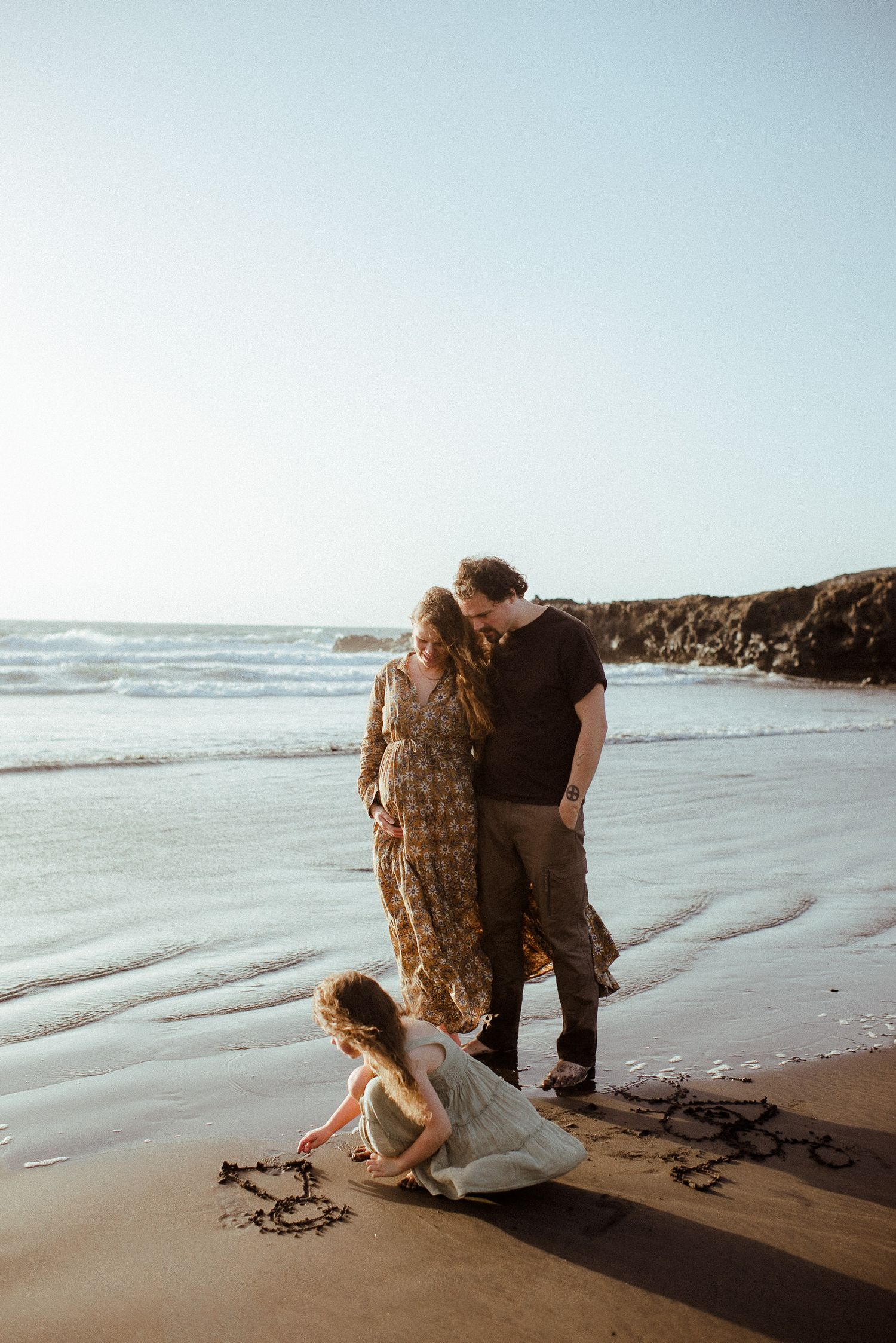 Family of three enjoying a beach day, parents watching their child draw in the sand by the ocean waves.