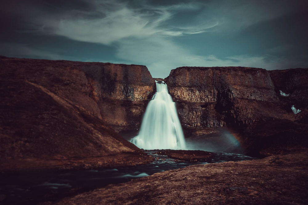 A dramatic waterfall cascades through rugged rocky cliffs under a moody dark sky in Iceland.
