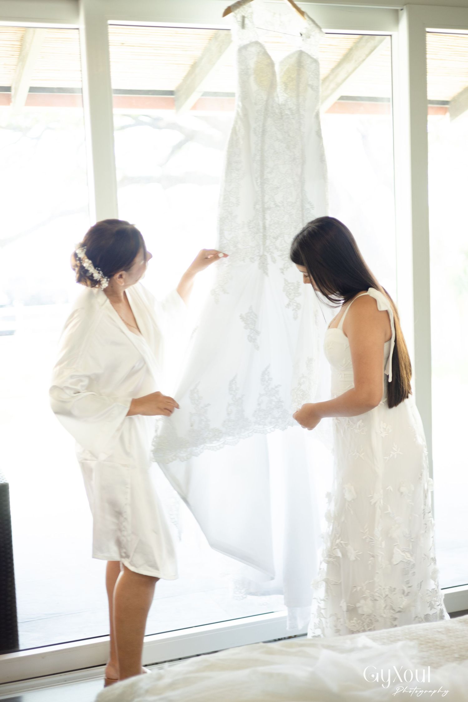 Two women in white attire admire a lace wedding gown hanging by a bright window on a wedding day.