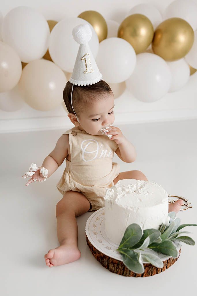 Baby in beige romper and number 1 party hat enjoys first birthday cake smash with gold and white balloon backdrop.