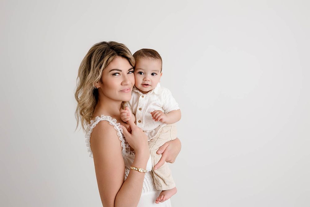 A young mother in a white lace dress holds her smiling baby boy in a bright studio setting by Temecula family photographer.