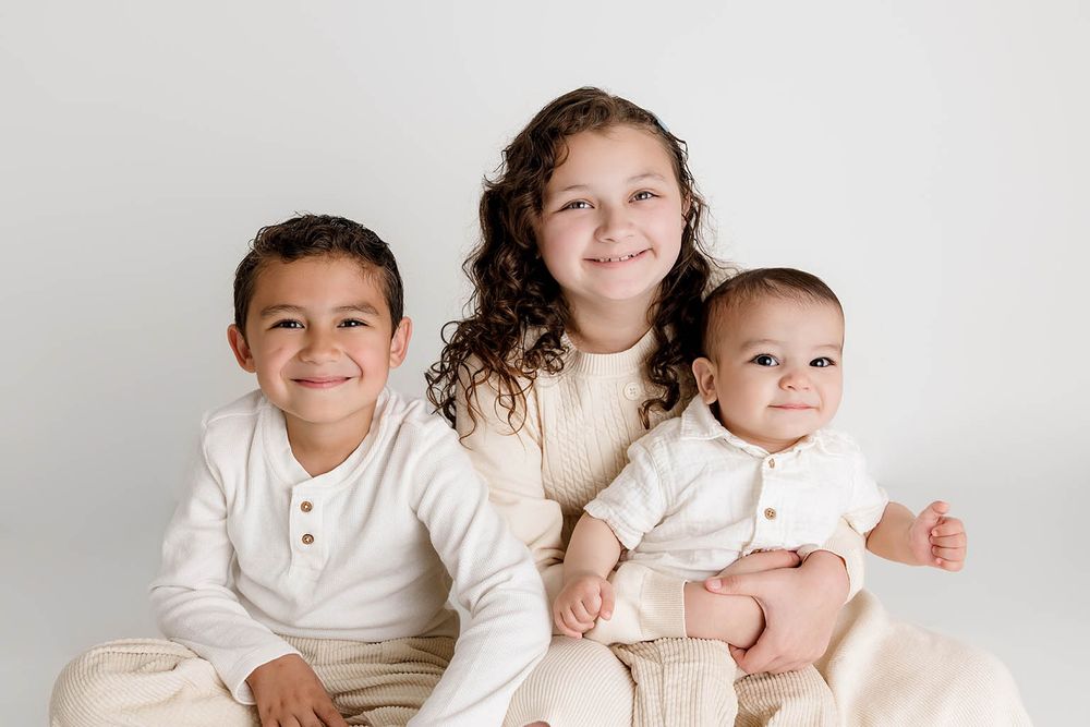 Three siblings in matching cream outfits pose together smiling for a professional studio portrait photo by Temecula family photographer.