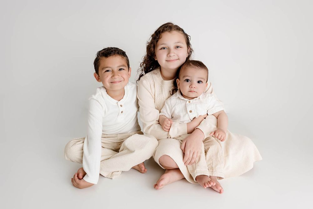 Three young siblings in matching cream outfits pose together on a white background by Temecula family photographer.