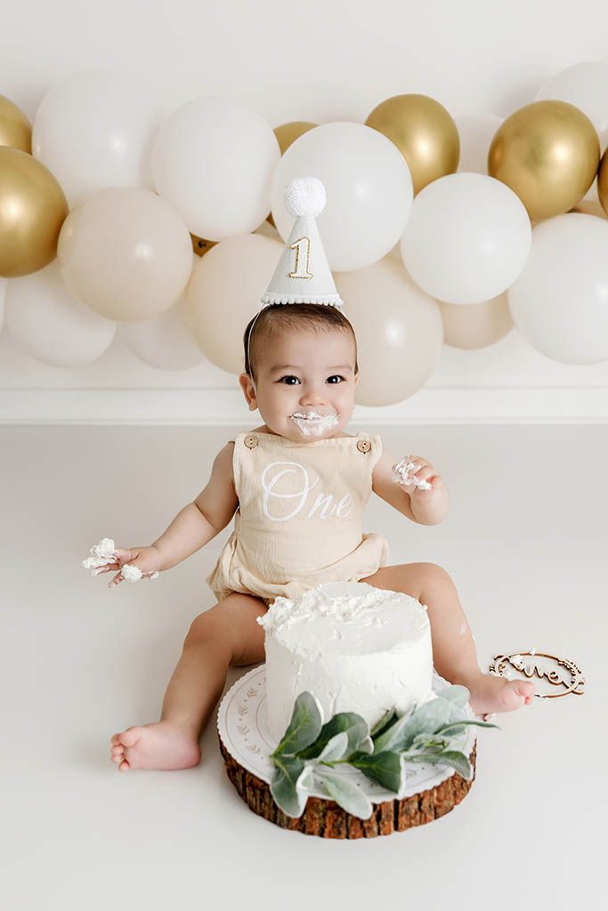 Baby's first birthday cake smash with gold and white balloons, party hat, and white frosted cake on wood slice.