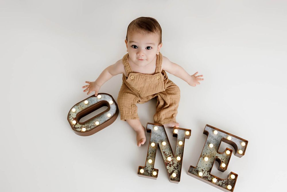 Baby in brown overalls lying next to marquee letters spelling ONE for first birthday photo.