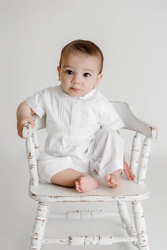 Sweet baby boy dressed in white outfit posed in a rustic white wooden chair.