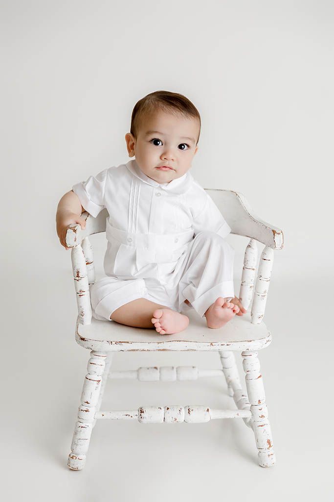 Adorable baby in white romper sitting in a vintage distressed white wooden chair.