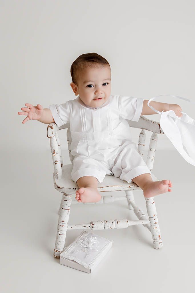 Baby in white romper sitting on a distressed white wooden chair with a gift box on the floor.