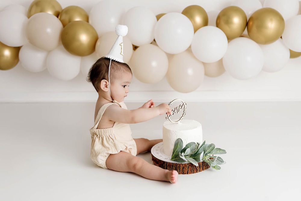 Baby in cream romper and party hat touches first birthday cake, surrounded by gold and white balloon garland backdrop.