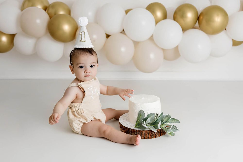 Baby in cream romper and party hat sits beside white cake with gold and white balloon garland backdrop.