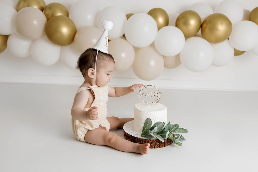 Baby in cream romper touches first birthday cake at elegant white and gold balloon backdrop photo shoot.