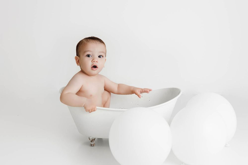 Adorable baby sitting in a white clawfoot bathtub surrounded by large white balloons on a white background.