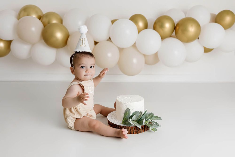 Baby in white onesie sits beside a white cake surrounded by gold and white balloons during a first birthday cake smash.