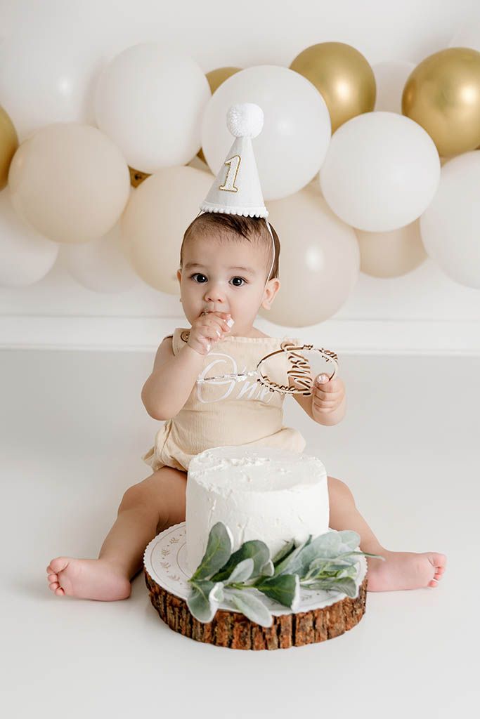 Adorable baby wearing number 1 party hat enjoys first birthday cake smash with gold balloon backdrop.