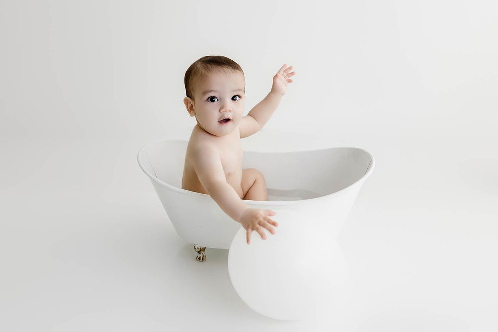 Cute baby sitting in a white mini clawfoot bathtub against a white background.