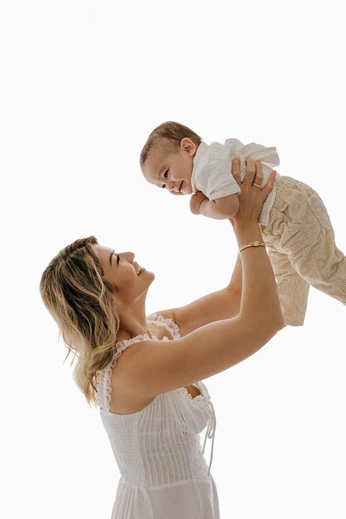 A joyful mother in a white dress lifts her smiling baby overhead against a bright white background by Temecula family photographer.