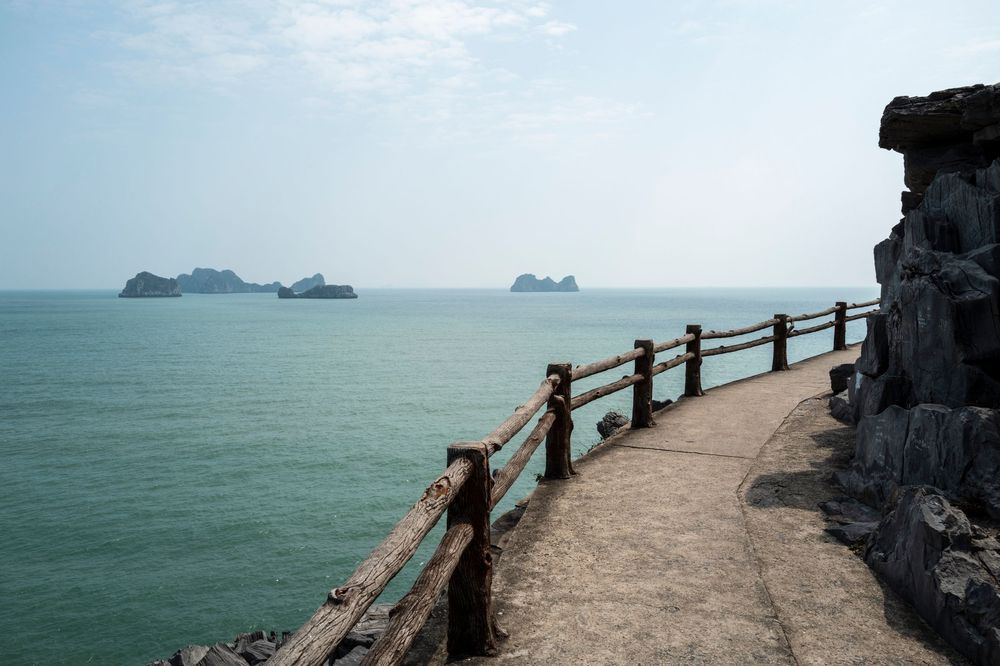 A scenic coastal walkway with wooden railings overlooking calm turquoise waters and distant rocky islands under a hazy sky.