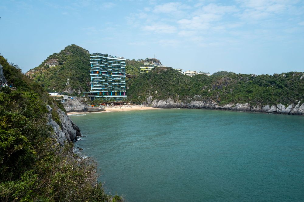 Scenic bay with turquoise waters, sandy beach, lush green hills, and a modern resort building in a tropical coastal setting.