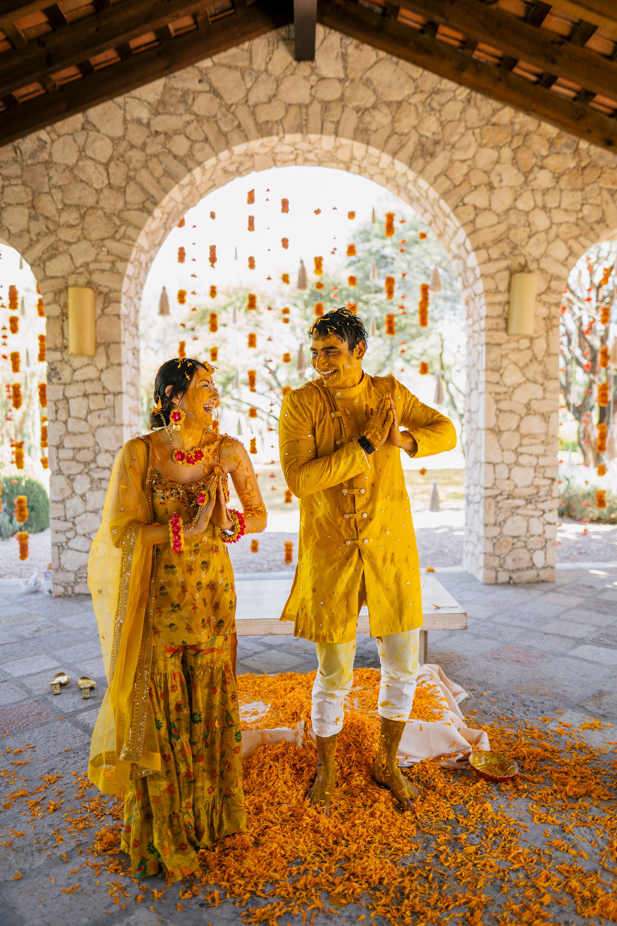 Indian couple in yellow outfits during Haldi ceremony, standing under stone arches with marigold flowers.